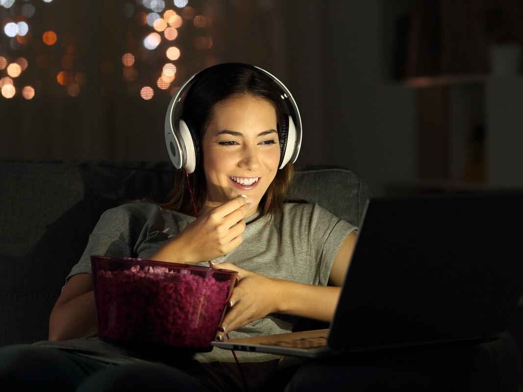 Frau mit Kopfhörern sitzt lächelnd mit Popcorn vor dem Bildschirm