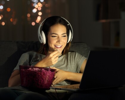 Frau mit Kopfhörern sitzt lächelnd mit Popcorn vor dem Bildschirm